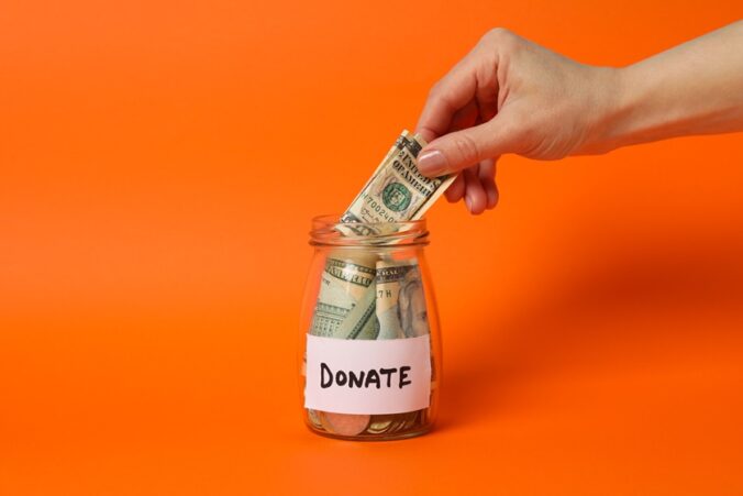 Female hand puts money in glass jar on orange background